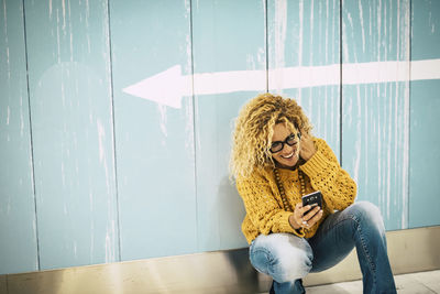 Mature woman using smart phone crouching by arrow symbol on wall