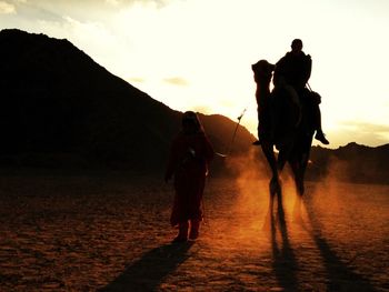 Woman walking with man on camel at desert against sky during sunset