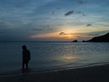 Rear view of silhouette boy standing on beach against sky during sunset