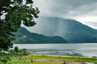 People on grass by lake and mountain against sky