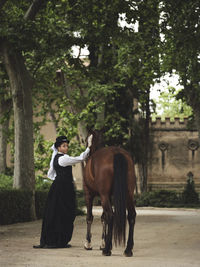 Full body of confident african american adult lady in elegant clothes and hat standing with brown horse while looking at camera near trees and building in daytime