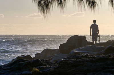 Rear view of silhouette man looking at sea while standing on rock against sky during sunset