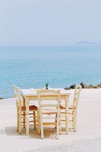 Light colored chairs and table with cactus by seaside against clear sky