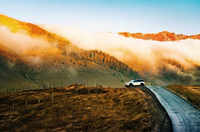 Car on road amidst land against misty mountain and sky at sunrise during autumn