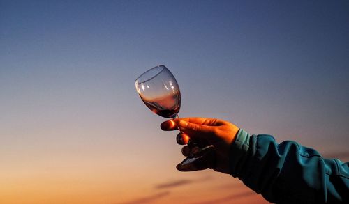 Cropped hand of man holding red wine in glass against sky during sunset