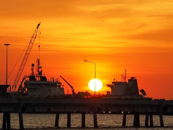 Silhouette cranes at harbor against orange sky
