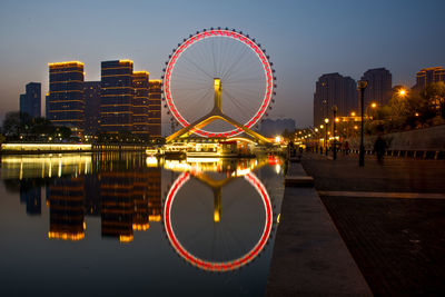 Illuminated ferris wheel by river against buildings at night
