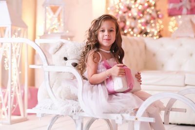 A beatiful girl with curly hair in white dress is holding of christmas box gift and sitting on sled 