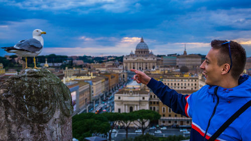 Young man pointing towards seagull while standing against cloudy sky in city during sunset