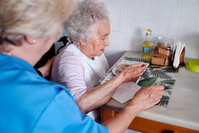 Cropped unrecognizable nurse in uniform near aged woman in wheelchair eating tasty soup at table with food in light kitchen at home