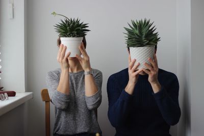 Couple holding potted plant while sitting on chair against wall at home