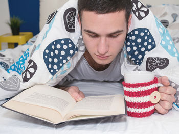 Close-up of young man reading book while holding cup on bed