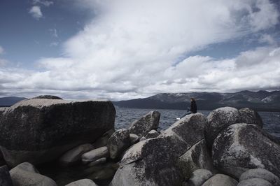 Side view of woman sitting on rocks by lake against cloudy sky