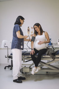 Full length of female doctor checking blood pressure of pregnant woman sitting on gurney in medical clinic