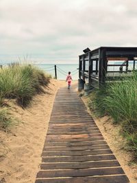 Rear view of girl walking on boardwalk at beach against cloudy sky