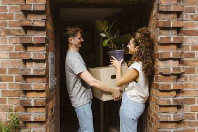 Happy couple with cardboard box and potted plant talking to each other at doorway