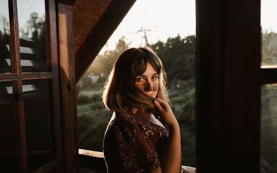 Portrait of young woman on ledge of wooden cabin at sunset looking back