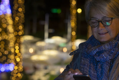 Smiling senior woman using mobile phone while standing against illuminated lighting equipment