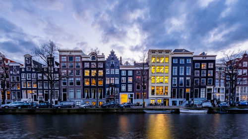 Buildings in city by river against cloudy sky