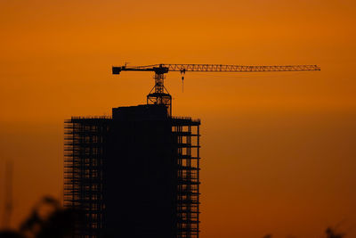 Low angle view of silhouette crane by building against sky during sunset