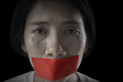 Portrait of crying woman mouth sealed with red tape against black background