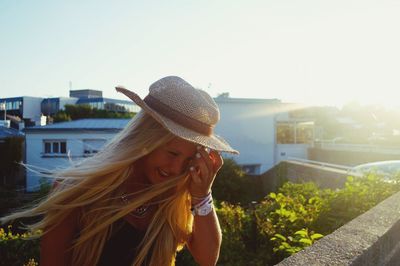 Smiling young woman wearing hat standing on building terrace against clear sky during sunny day