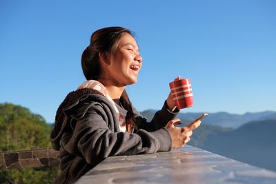 Smiling woman using mobile phone while having coffee against clear blue sky