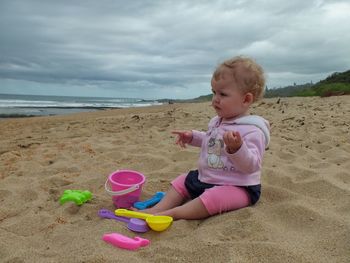 Full length of girl sitting by toys at sandy beach against cloudy sky