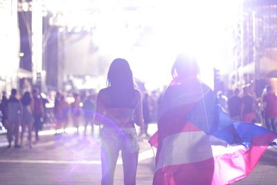 Rear view of female friends with puerto rican flag standing on illuminated street at night