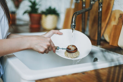 Young woman washes dishes with wooden brush with natural bristles at window in the kitchen