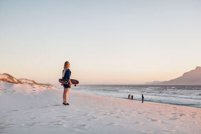 Side view of young woman with skateboard standing at beach against clear sky during sunset