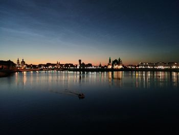 Illuminated buildings by river against sky during sunset