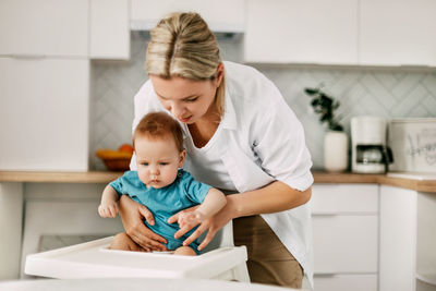 A young mother puts her young son in a baby high chair for feeding