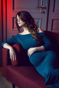 Pregnant woman in blue dress lying on red sofa in studio