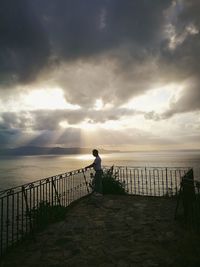 Man standing on railing by sea against sky during sunset