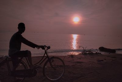 Silhouette man on bicycle at beach against sky during sunset