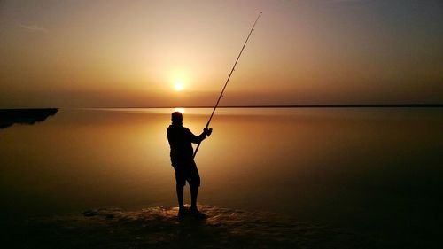 Silhouette man fishing in sea against sunset sky