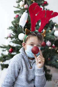 Portrait of boy wearing mask sitting against christmas tree at home