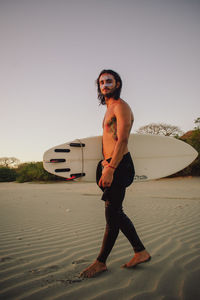 Portrait of young man with surfboard standing on beach against clear sky