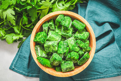 Frozen spinach cubes in a wooden bowl with fresh parsley and green onions on a light gray cement .