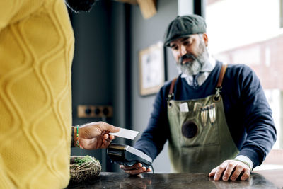 Crop male customer making payment for hairstyle with plastic card on nfc terminal in hands of barber in grooming salon