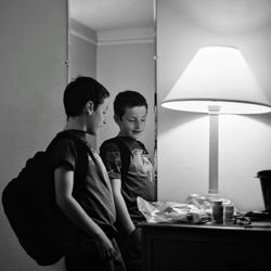 Side view of boy standing by mirror and electric lamp at home