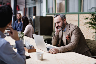 Tired businessman using laptop while sitting with colleagues at table in meeting