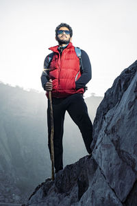 Young indian traveler standing confidently on top of the mountain over a cliff, wearing snow jacket.