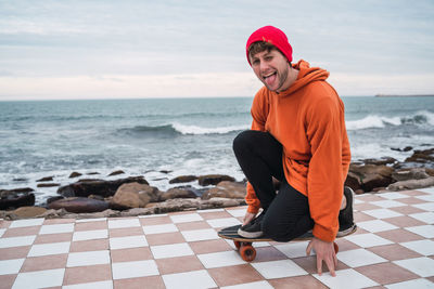 Full length of man sitting on skateboard by sea against sky