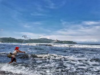 Man throwing fishing net in sea against sky