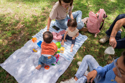 From above of small baby girl and boy sitting on plaid with toys near crop parents in park