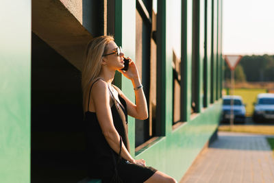 Stylish young woman walking around the city is resting sitting on the parking wall and talking