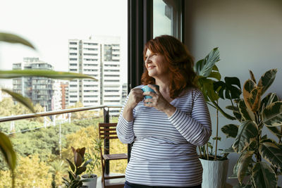 Smiling woman drinking coffee while standing by balcony at home