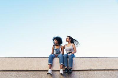 Female friends sitting on retaining wall against clear sky